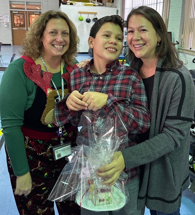 A Children’s Education Center teacher, a student, and the student's mother who is holding the gingerbread house they decorated together.