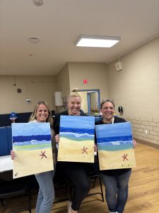 Three attendees of Round HillRoad's Paint Night hold up their paintings of the ocean